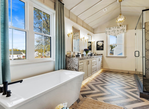 Spacious modern bathroom with a freestanding tub, double-sink vanity, large glass shower, chevron-patterned floor, blue curtains, and large windows letting in natural light.