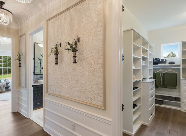 A spacious modern walk-in closet with white shelving, drawers, and hanging rods, connected to a bright bedroom and bathroom. The closet features organized clothing and large windows for natural light.