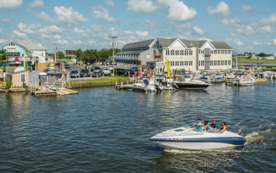 A speedboat with several people rides on a river near a marina with docked boats. Buildings, cars, and people are visible onshore under a blue sky with scattered clouds.