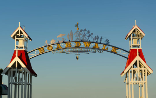 An arched entrance sign reading Boardwalk Ocean City, Maryland with decorative silhouettes of people and rides, flanked by two white towers with red roofs against a clear blue sky.