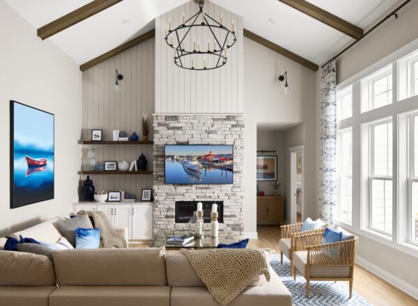 Bright living room with high ceiling, large windows, beige sectional sofa, wicker chair, and a stone fireplace. Nautical-themed decor, blue accent pillows, and wall art featuring boats add to the airy, cozy atmosphere.