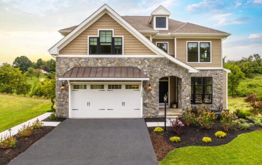 Two-story house with stone and tan siding, black-framed windows, a white double garage, and a manicured garden in front, set against a green lawn and partly cloudy sky.