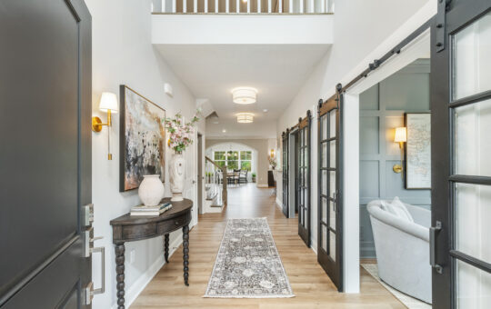 Bright entryway with light wood floors, a patterned runner rug, a round table with decor, artwork on the wall, and double glass doors opening to a cozy sitting room. A staircase and dining area are visible in the background.