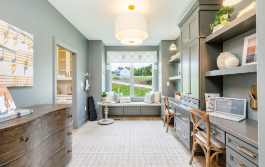 A bright home office with gray walls, built-in desks, wooden chairs, a large window with a window seat, and shelves displaying books, plants, and decor. Light fixture hangs from the ceiling; outdoor scenery is visible.