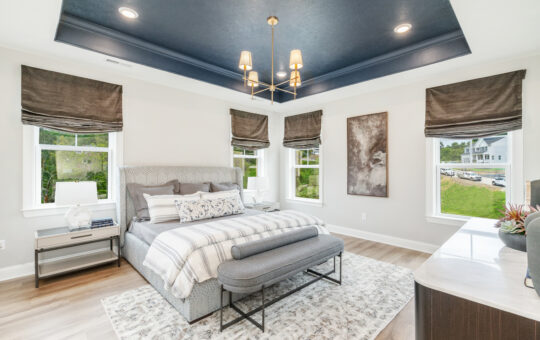 Modern bedroom with a large bed, gray bench, and patterned rug. Four windows with dark roman shades let in natural light. A navy blue ceiling features a contemporary chandelier. Theres abstract wall art and neutral décor.