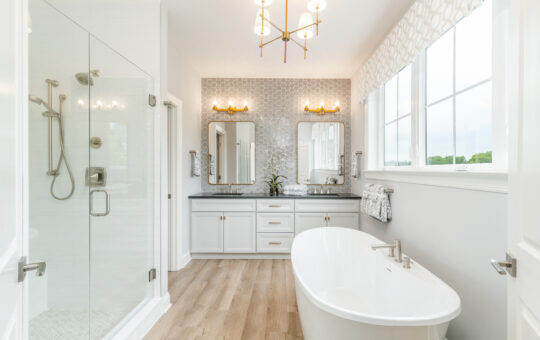 Modern white bathroom with a freestanding bathtub, glass-enclosed shower, dual sinks with mirrors, patterned accent wall, gold light fixtures, and large windows letting in natural light.