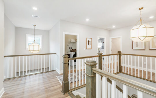 Bright upstairs hallway with wooden floors, white railings, modern geometric pendant lights, and multiple doorways leading to different rooms in a home. Natural light comes in through a window.