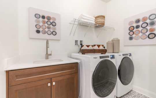 A modern laundry room with a front-loading washer and dryer, a wooden cabinet with a sink, floating wire shelf with towels and baskets, and abstract dot artwork on white walls.