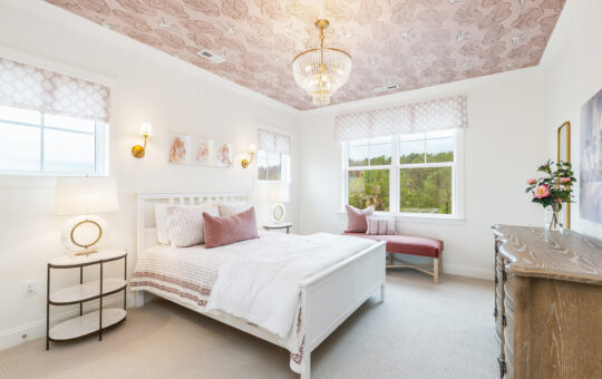Bright bedroom with a white bed, pink and white bedding, floral ceiling, chandelier, wooden dresser, round bedside table with lamp, two windows, pink bench, wall art, and fresh flowers in a vase.