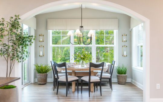 A bright dining area with large windows, a wooden table set for six, cushioned chairs, a black chandelier, and green plants in woven baskets by the windows. Sunlight fills the room, highlighting the greenery outside.