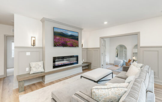 A bright living room with a large sectional sofa, patterned pillows, a bench, wall-mounted TV above an electric fireplace, and neutral decor. An adjacent room with chairs is visible through an open doorway.