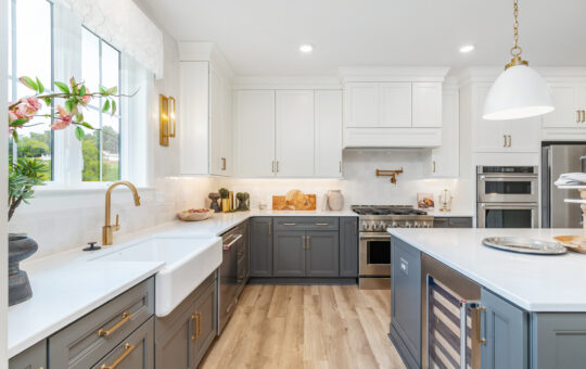 Modern kitchen with white upper cabinets, grey lower cabinets, a farmhouse sink, gold fixtures, stainless steel appliances, a kitchen island with a wine fridge, and wood flooring. Sunlight streams in from a large window.