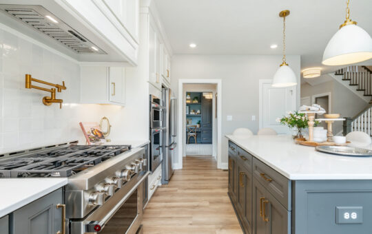 Modern kitchen with stainless steel gas stove, white cabinets with gold handles, large gray island with white countertop, pendant lights, light wood flooring, and a view into an adjoining pantry.