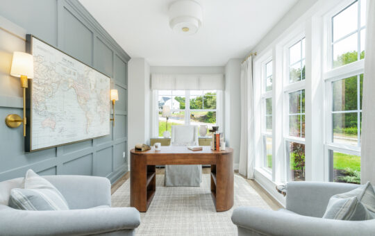 Bright home office with large windows, a wooden desk, two gray armchairs, a world map on a paneled wall, and neutral decor. Sunlight streams in, highlighting the airy, organized space with a garden view outside.