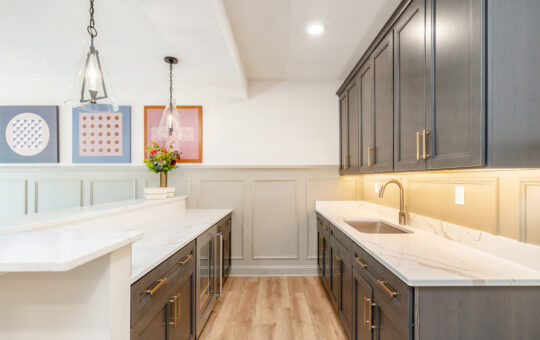 Modern kitchen with dark cabinets, brass handles, white marble countertops, wood flooring, pendant lights, a vase of flowers on the counter, and abstract art on the wall. Under-cabinet lighting illuminates the workspace.