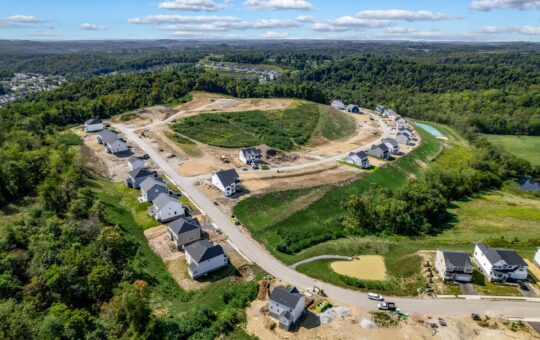Aerial view of a suburban housing development under construction, with new homes lining curved roads, surrounded by green trees and hills under a blue sky with scattered clouds.