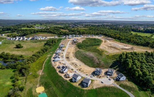 Aerial view of a suburban neighborhood under construction, with several completed houses, partially built homes, construction equipment, green fields, trees, and a small pond under a bright blue sky with clouds.
