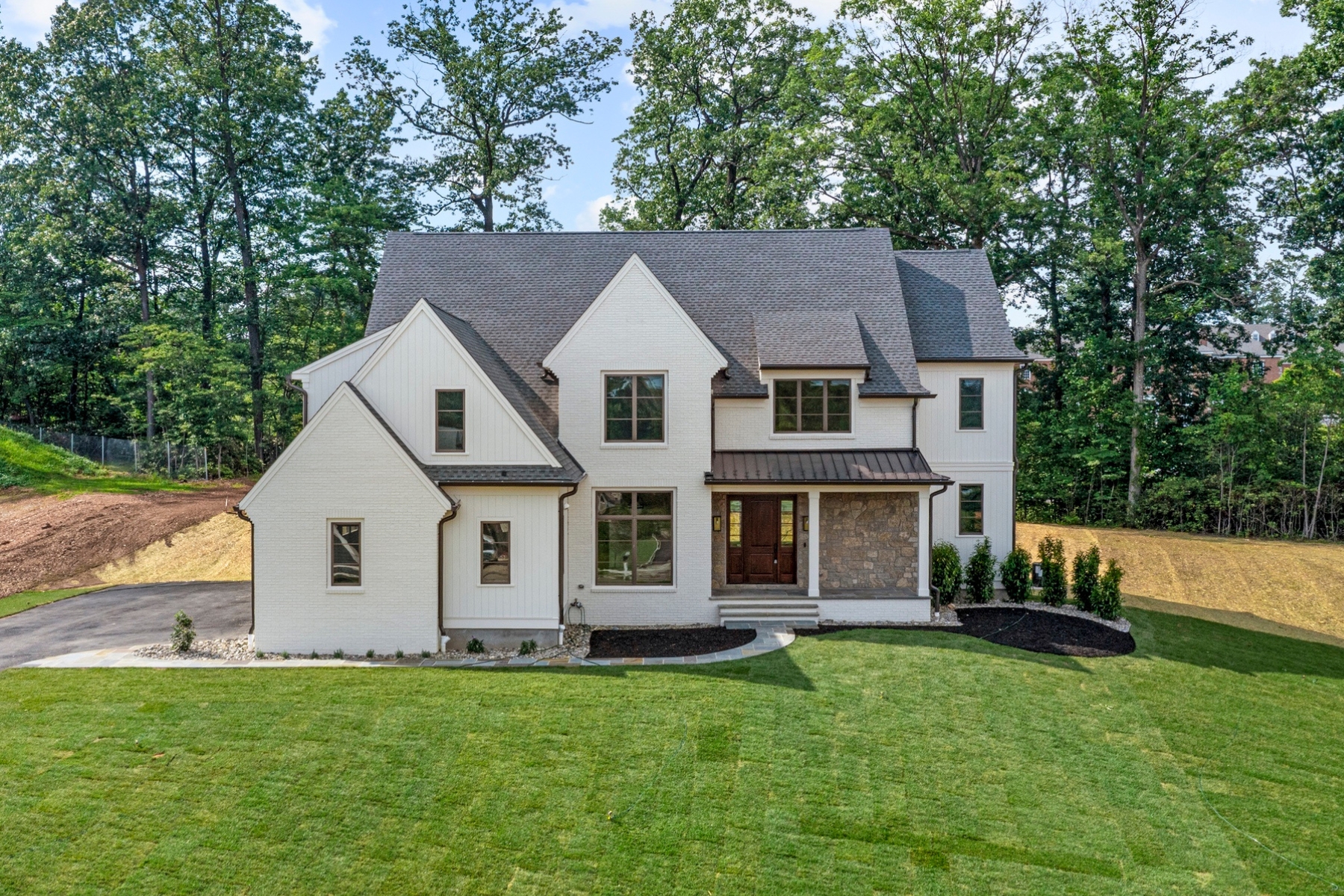 A two-story modern house with light brick and stone exterior, multiple large windows, and a covered front porch, situated on a spacious green lawn bordered by trees—exemplifying the elegance of North Wayne luxury homes.