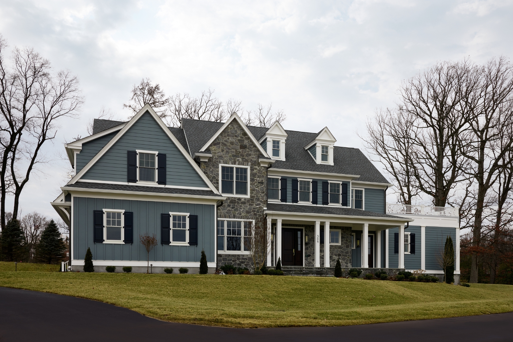 A large, two-story suburban house with blue siding, white trim, and stone accents sits on a well-manicured lawn with leafless trees and a paved driveway—an ideal example of the elegance found in North Wayne luxury homes.