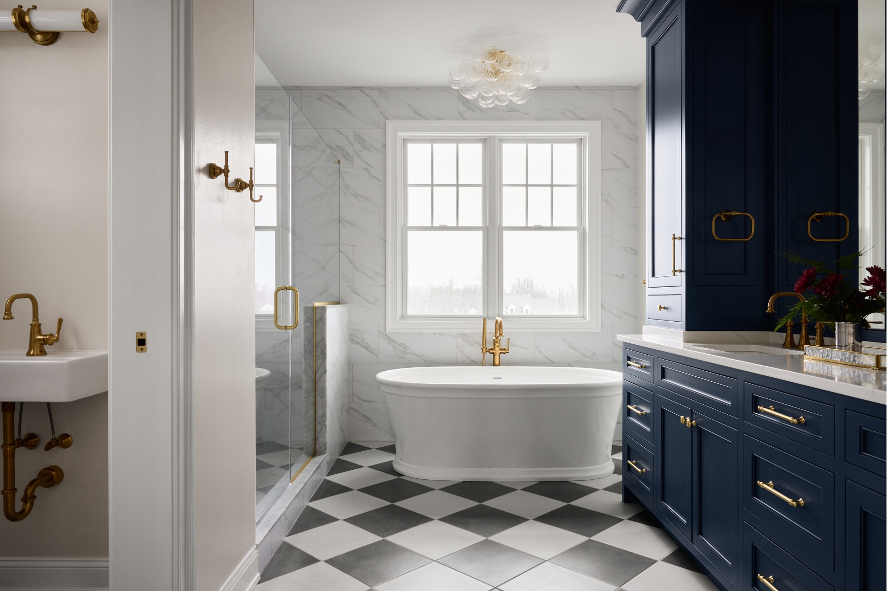 Elegant bathroom in North Wayne luxury homes with a freestanding tub, gold fixtures, checkered black-and-white floor, marble walls, large windows, blue cabinetry, and a glass shower enclosure.