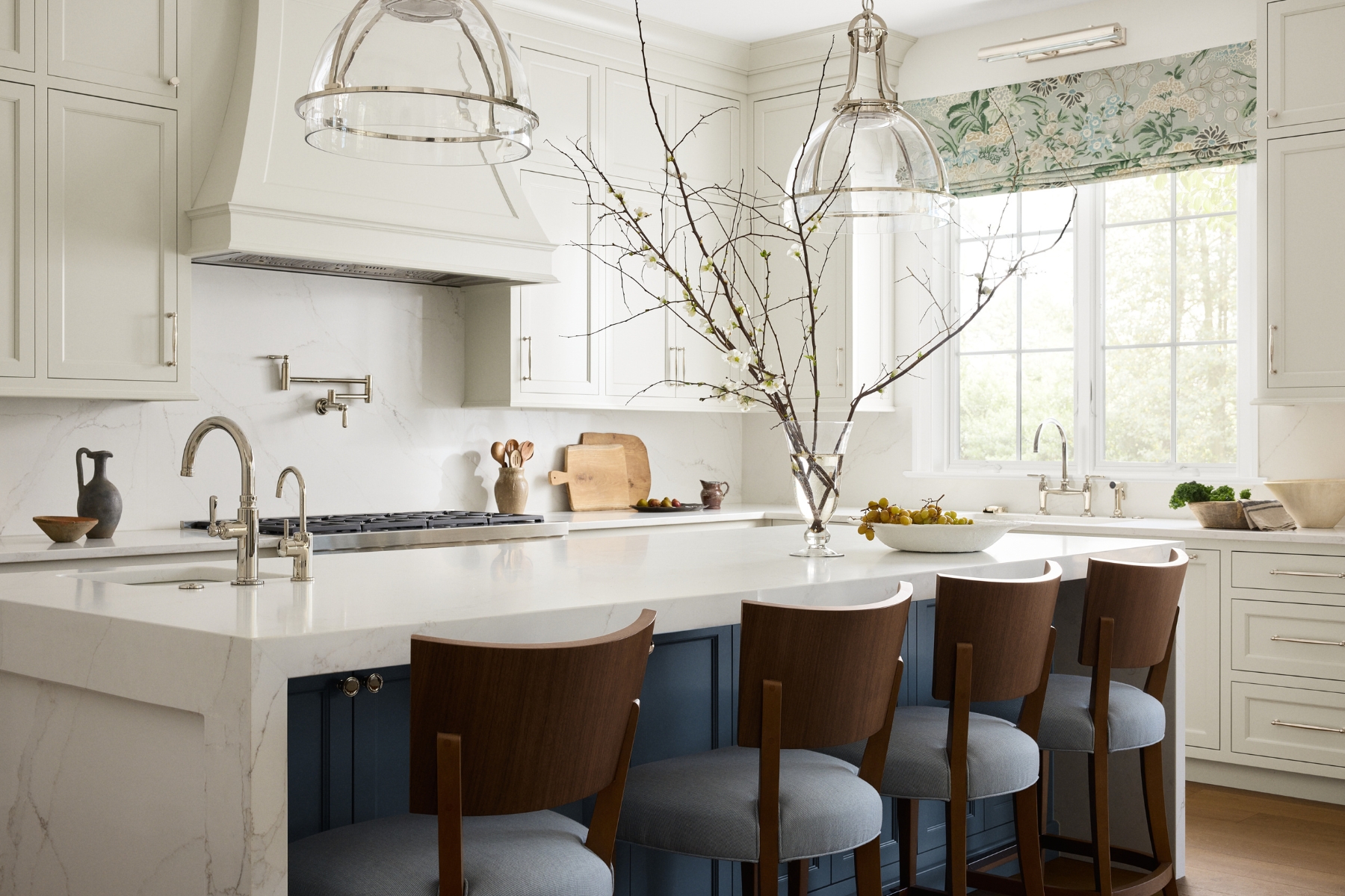 A bright, modern kitchen in North Wayne homes with white cabinets, marble countertops, a large island with wooden chairs, pendant lights, and a branch centerpiece. Sunlight streams through a window with a floral shade above the farmhouse sink.