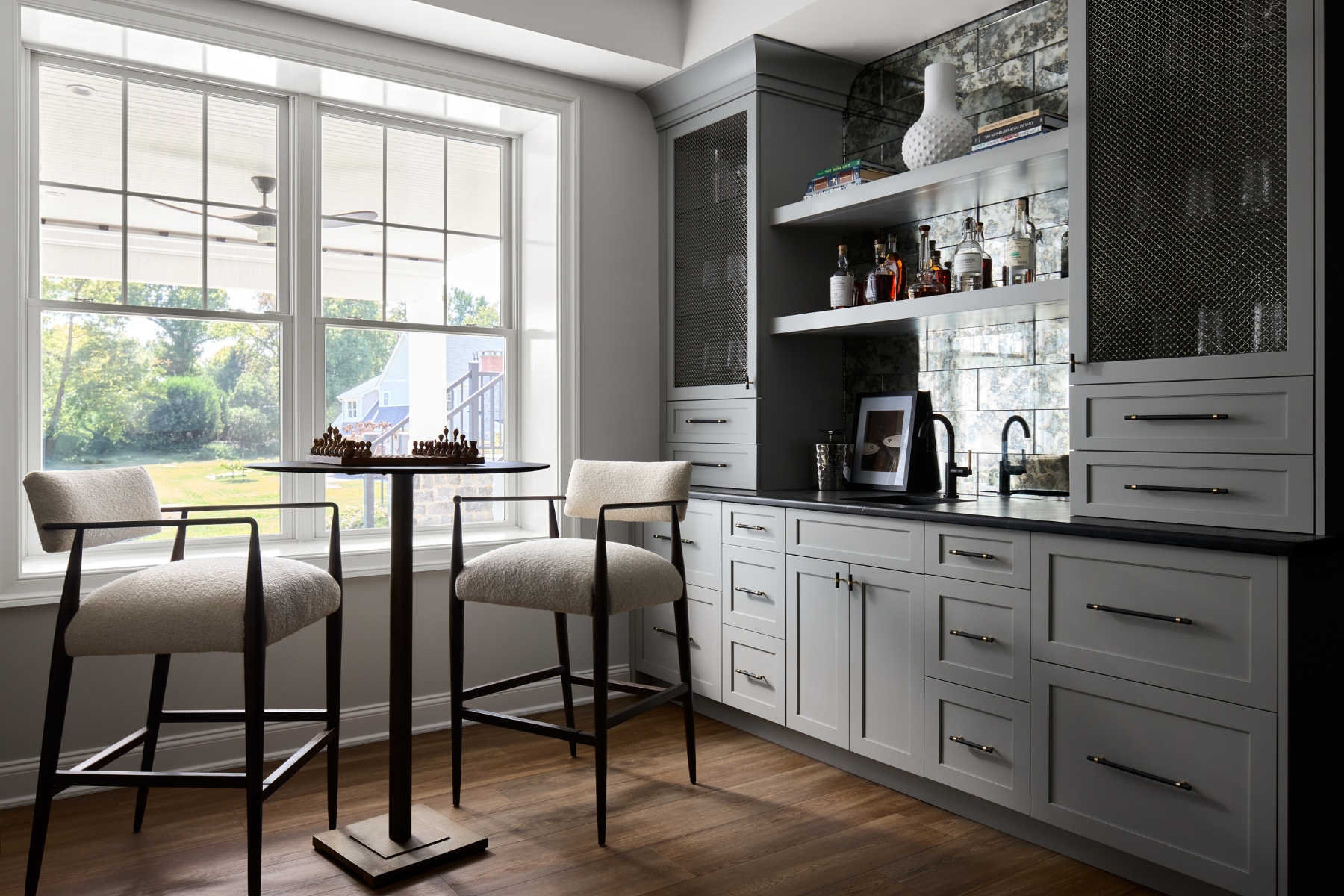 A bright home bar area found in many North Wayne luxury homes features a black countertop, glass shelves, white cabinets, a small sink, and a square table with cushioned chairs by large windows overlooking a green yard.