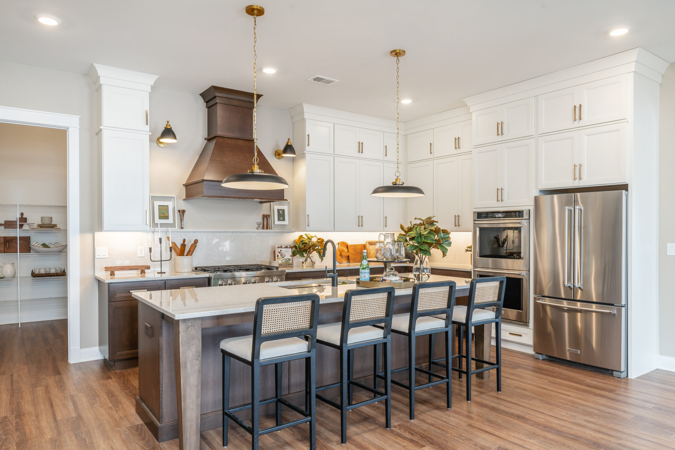 Modern kitchen with white cabinets, stainless steel appliances, a large island with four barstools, pendant lights, a wooden floor, and decorative plants and accessories on the counters.