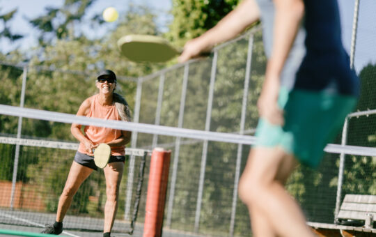 Two people play pickleball on an outdoor court. A woman in an orange tank top and black shorts prepares to hit the ball, while another player in a gray shirt and green shorts stands in the foreground.
