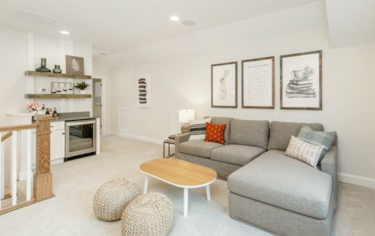 A modern living room with a gray sectional sofa, two poufs, a wooden coffee table, and framed wall art. There is a small bar area with a mini fridge and shelves, and neutral decor throughout the space.