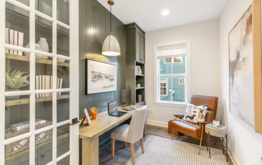 A modern home office with a wooden desk, laptop, stationery, and artwork on green paneled walls. A brown leather chair and small table are placed near a window with a view of houses outside. Shelves hold books and plants.