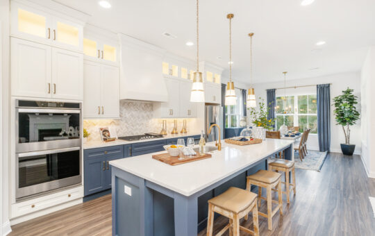 Modern kitchen with white and navy cabinets, a large island with stools, pendant lights, built-in ovens, gas stove, and a dining area with large windows and greenery in the background.