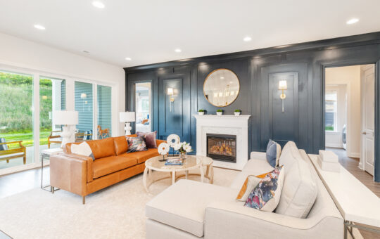 Modern living room with a tan leather sofa, light beige sectional, round coffee table, and white fireplace set in a dark accent wall with a large round mirror and sconces. Floor-to-ceiling windows overlook a patio and greenery.