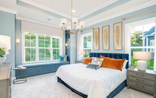 Bright, modern bedroom with blue walls, a large bed with white linens and orange pillows, a window seat, two big windows, framed artwork above the bed, and a gold chandelier on the ceiling.