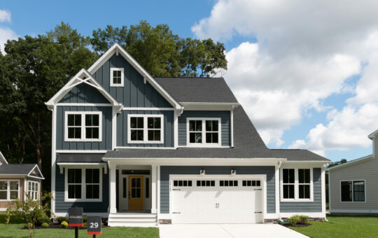 A modern two-story blue house with white trim, large windows, a covered front porch, and a double garage sits on a neatly manicured lawn under a partly cloudy sky. A “For Sale” sign is visible in the yard.