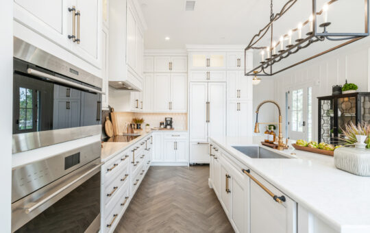 Modern, bright kitchen with white cabinets, stainless steel appliances, a large island with a farmhouse sink, gold fixtures, and herringbone wood flooring. Natural light enters through glass doors.