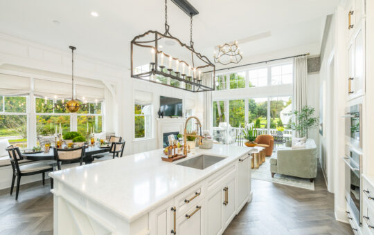 Bright, modern kitchen and living area with white cabinets, large island, dining table, cozy seating, and floor-to-ceiling windows overlooking a green outdoor space. Natural light fills the open, airy room.