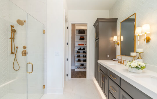 Modern bathroom with a glass shower featuring gold fixtures, double sink vanity with gold accents, white countertops, wall sconces, and a view into a closet with shelves for shoes and hats.