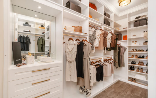 A modern, organized walk-in closet featuring neatly hung clothes, shelves with handbags and shoes, white cabinetry with gold handles, and a vanity area with a mirror and decorative items.