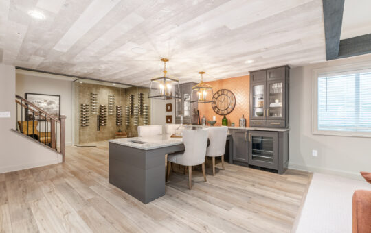 Modern basement with light wood flooring, a small dining area with gray cabinetry, a bar, elegant pendant lights, and a glass-enclosed wine rack along the back wall. A window lets in natural light from the right.