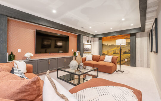 A modern basement living room with orange sectional sofas, a large wall-mounted TV, built-in cabinets, glass coffee table, and a scenic mural on the back wall, decorated with vases and framed photos.