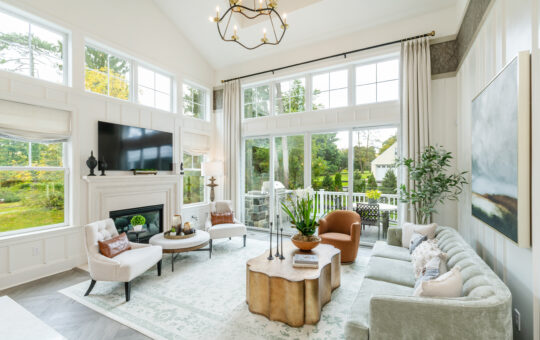 Bright, elegant living room with large windows, neutral-toned furniture, a cozy fireplace, TV, potted plants, and a modern chandelier. The space opens to a green outdoor patio with trees visible through the glass doors.
