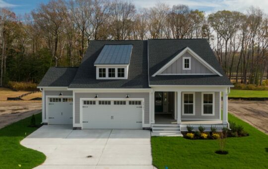 A modern two-story gray house with white trim, a covered front porch, two garage doors, and a manicured lawn. Leafless trees and a partly cloudy sky are visible in the background.