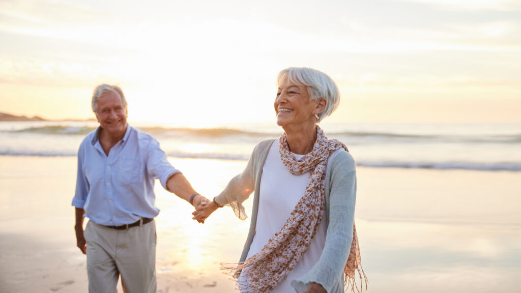 Laughing senior woman leading her husband by the hand while walking along a sandy beach at sunset in summer