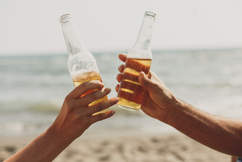 two people toasting glass beer bottles on the beach