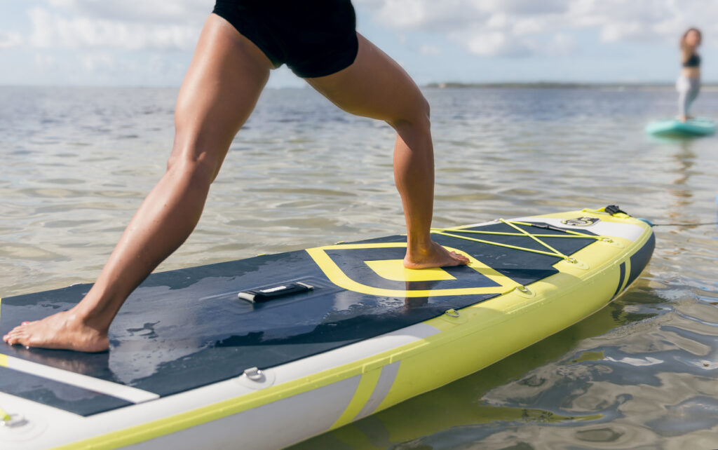 Cropped shot of a young woman doing warrior pose on top of a surf paddle board while paying attention to her yoga instructor in the distance.