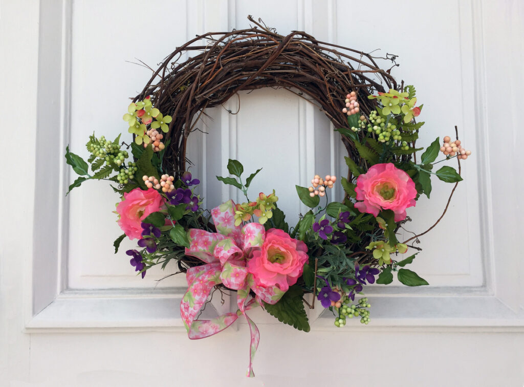 A beautiful wreath for spring on a white door with pink  and purple flowers and greenery.  It has a pink and green flower patterned bow on a white background.