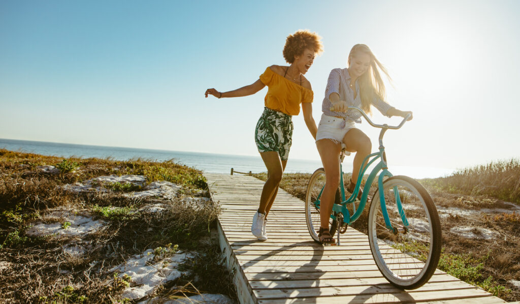 two friends riding bikes and walking on sunny beach boardwalk