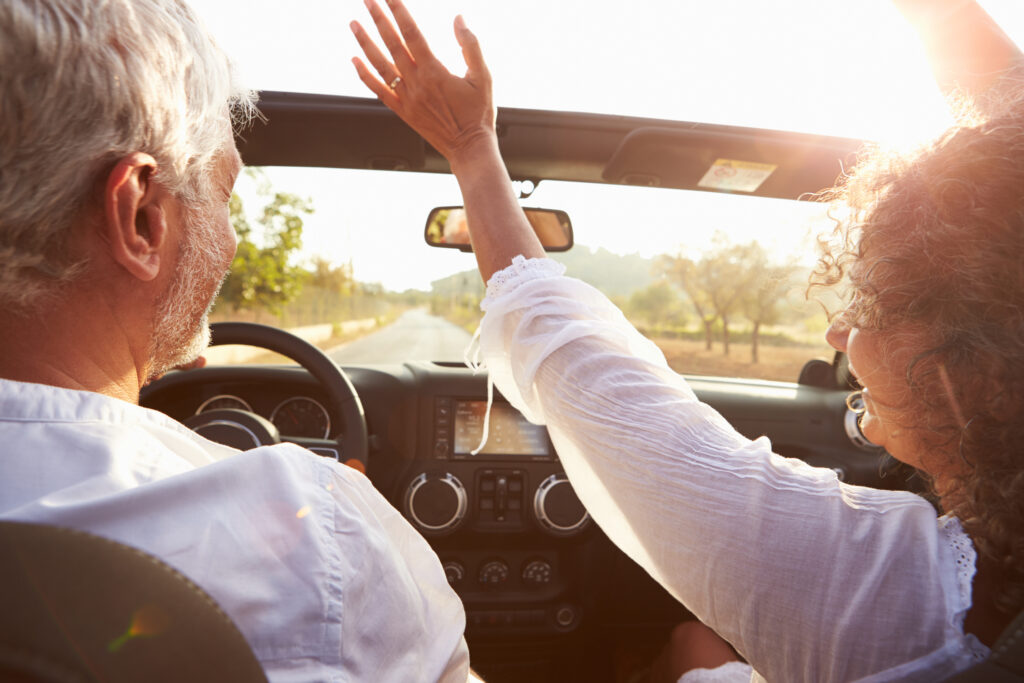 Mature Couple Driving Along Country Road In Open Top Car Hoding Arms Up
