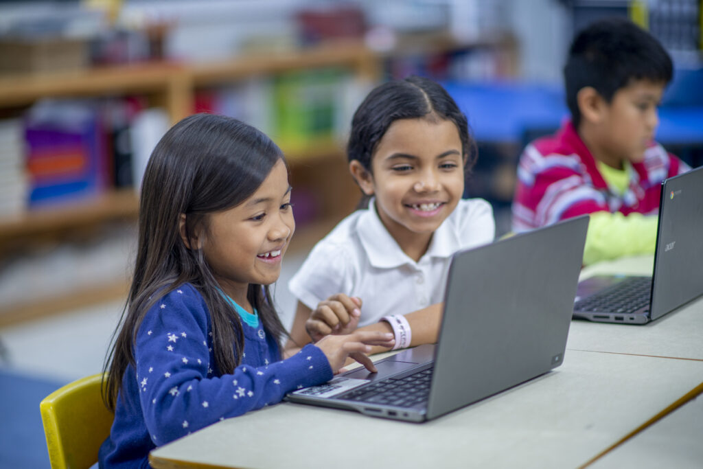 Two elementary students sit at a desk as they work together on a laptop during a computer lab.