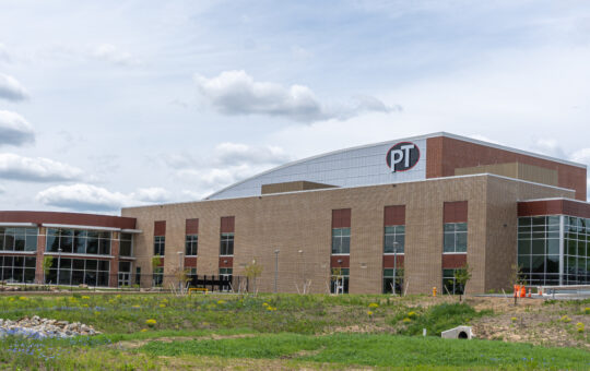 Modern two-story building with large windows and a brick exterior, featuring the letters PT on its corner. Theres a grassy area with wildflowers in the foreground and a cloudy sky above.